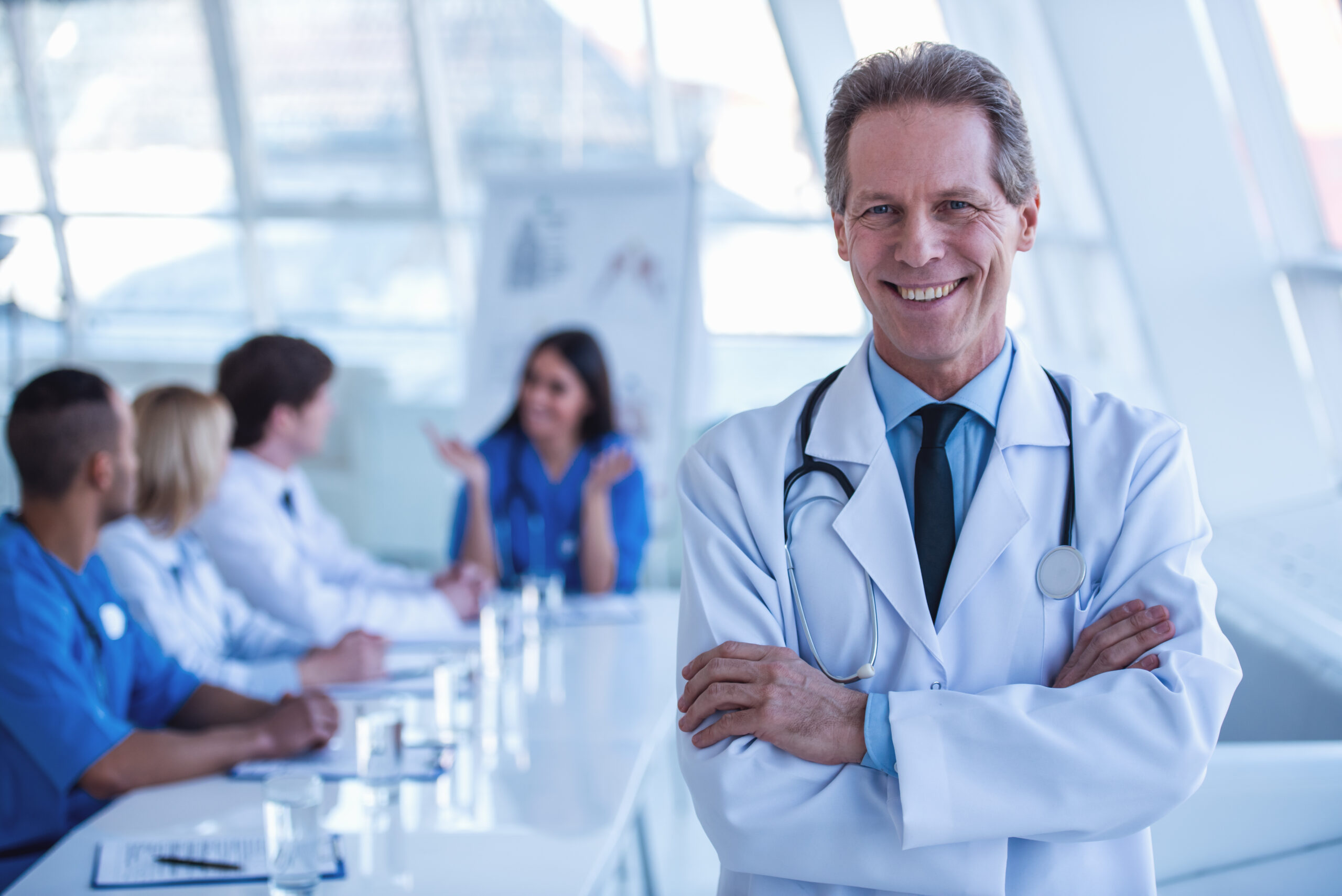 Handsome mature medical doctor is looking at camera and smiling while his colleagues are sitting in the background