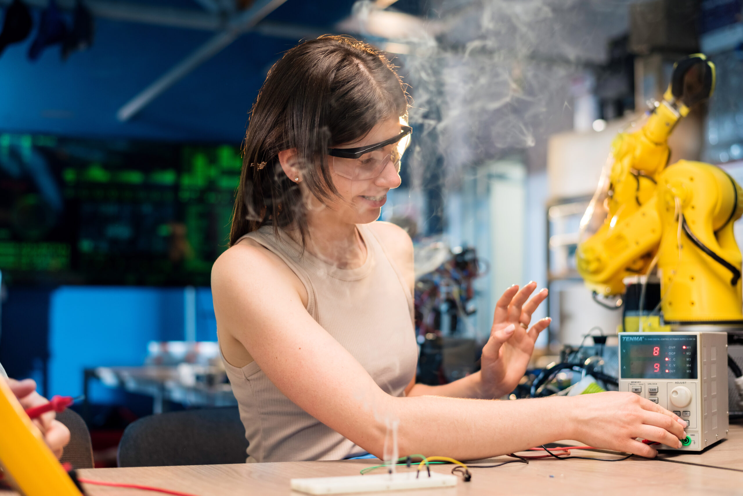 Young woman in protective glasses doing experiments with electricity in a laboratory. Smoke coming out from a plate, tools on the table